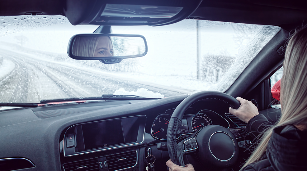 View through windshield of a car with a snowy scene, with female driver's eyes in mirror